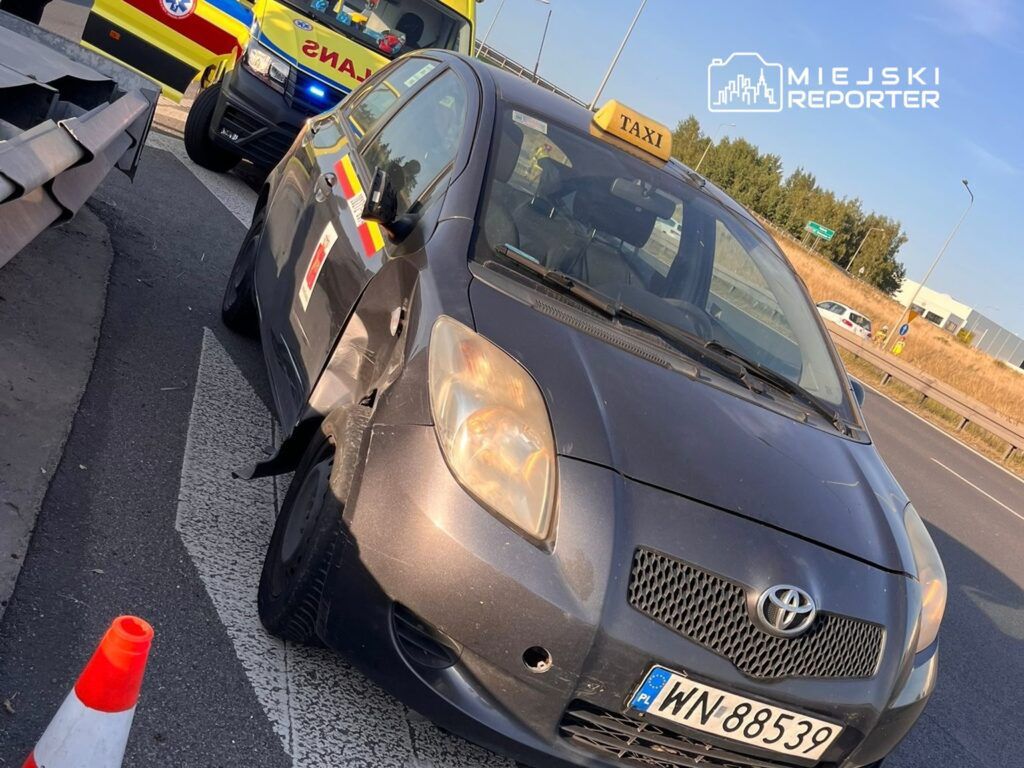 Czarny samochód osobowy z oznaczeniem taksówki uszkodzony na poboczu drogi, w tle karetka pogotowia.