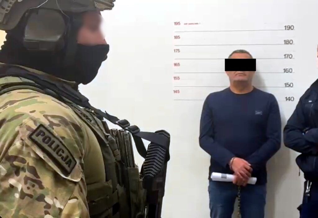 Policjant w kamizelce taktycznej stoi obok mężczyzny w kajdankach, który jest fotografowany na tle ściany z miarką.