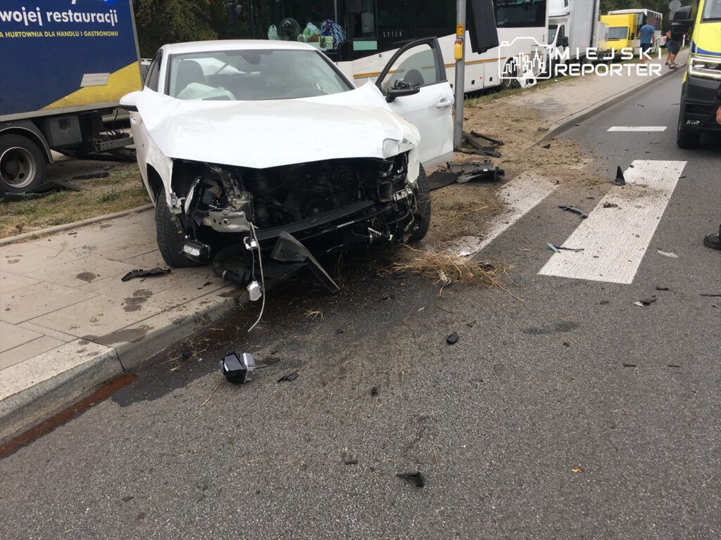 Uszkodzony biały samochód osobowy stoi przy krawężniku, obok leżą fragmenty pojazdu i zanieczyszczenia na drodze.