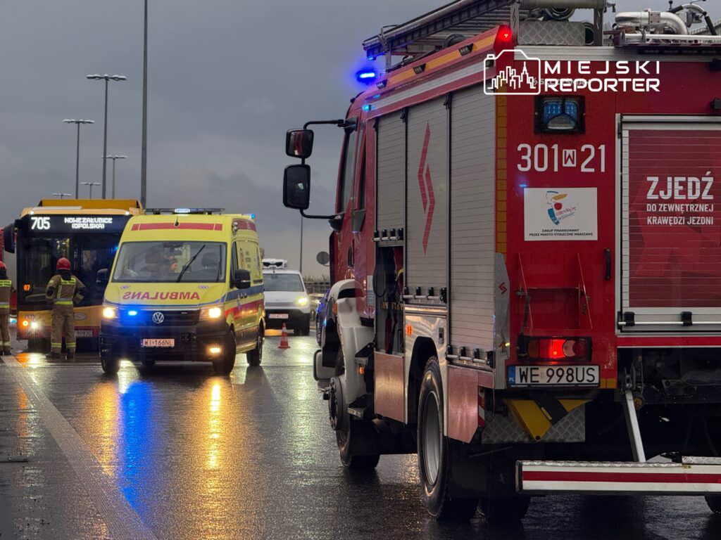 Zespół ratunkowy w akcji, ambulans i wóz strażacki na mokrej jezdni, w tle autobus komunikacji miejskiej.