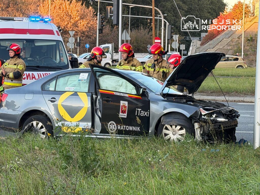 W pobliżu skrzyżowania, strażacy w hełmach rozmawiają obok uszkodzonego samochodu taxi z otwartą maską.