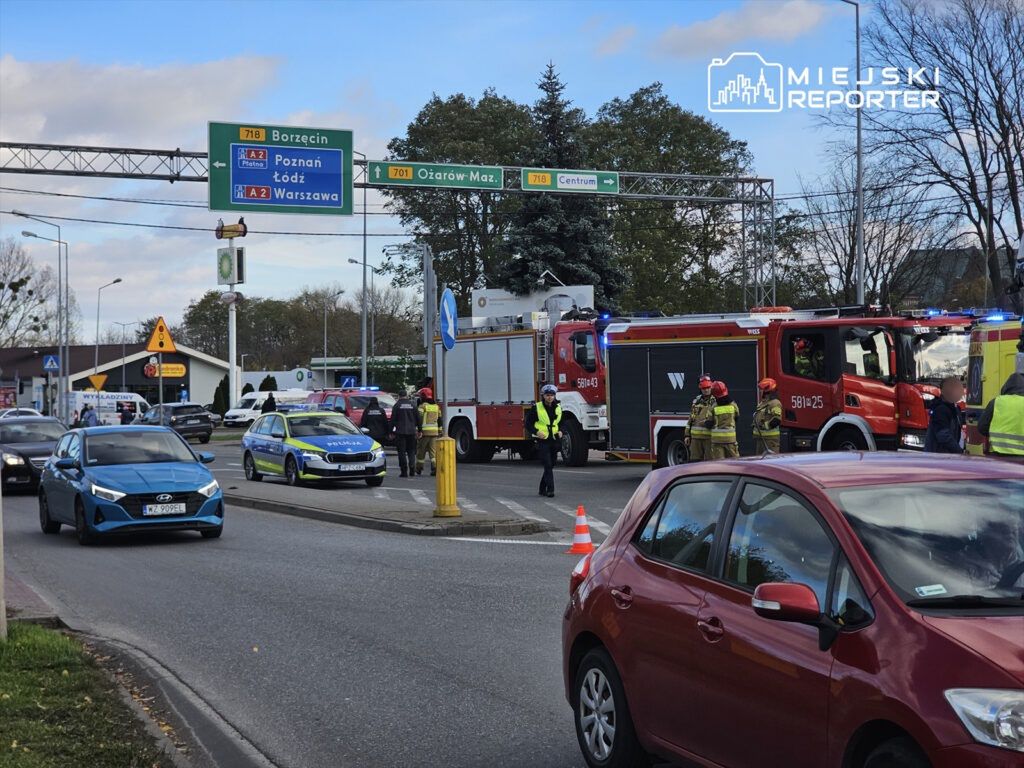 Na skrzyżowaniu ruchu drogowego stoją pojazdy ratunkowe, w tym wóz strażacki i radiowóz policji, a obok grupka strażaków.