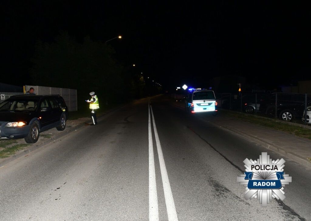 Policjant w odblaskowej kamizelce kontroluje dokumenty kierowcy przy zatrzymanym samochodzie na oświetlonej ulicy.