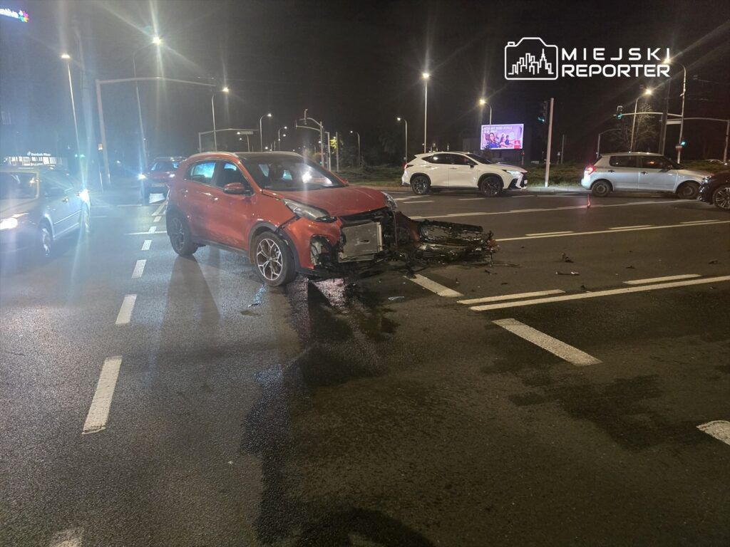 Czerwony samochód osobowy z uszkodzonym przodem stoi na skrzyżowaniu, wokół inne pojazdy i rozlane płyny.