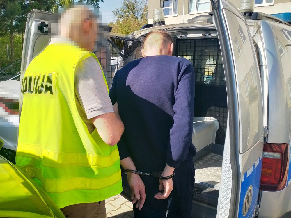 Mężczyzna w kajdankach wchodzi do policyjnego auta, obok stoi funkcjonariusz w kamizelce odblaskowej.