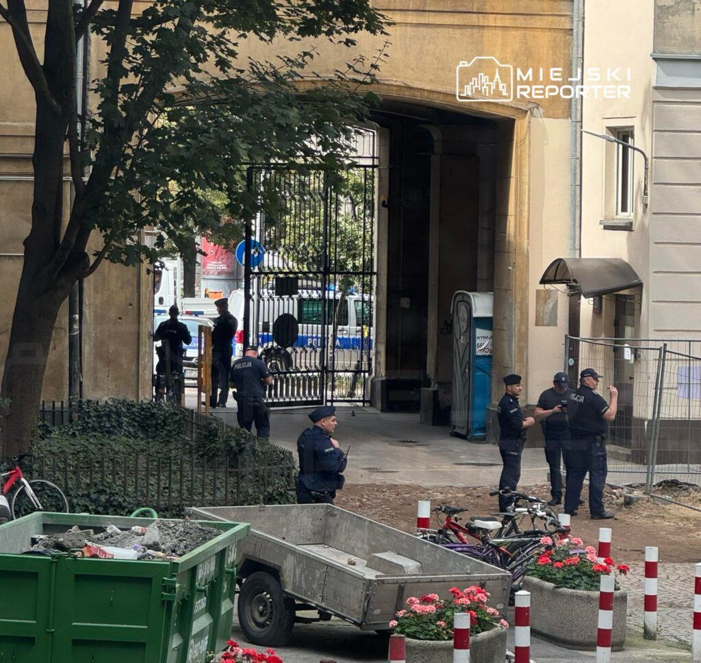 Grupa policjantów rozmawia w bramie budynku, obok stoi kontener na odpady oraz rowery.