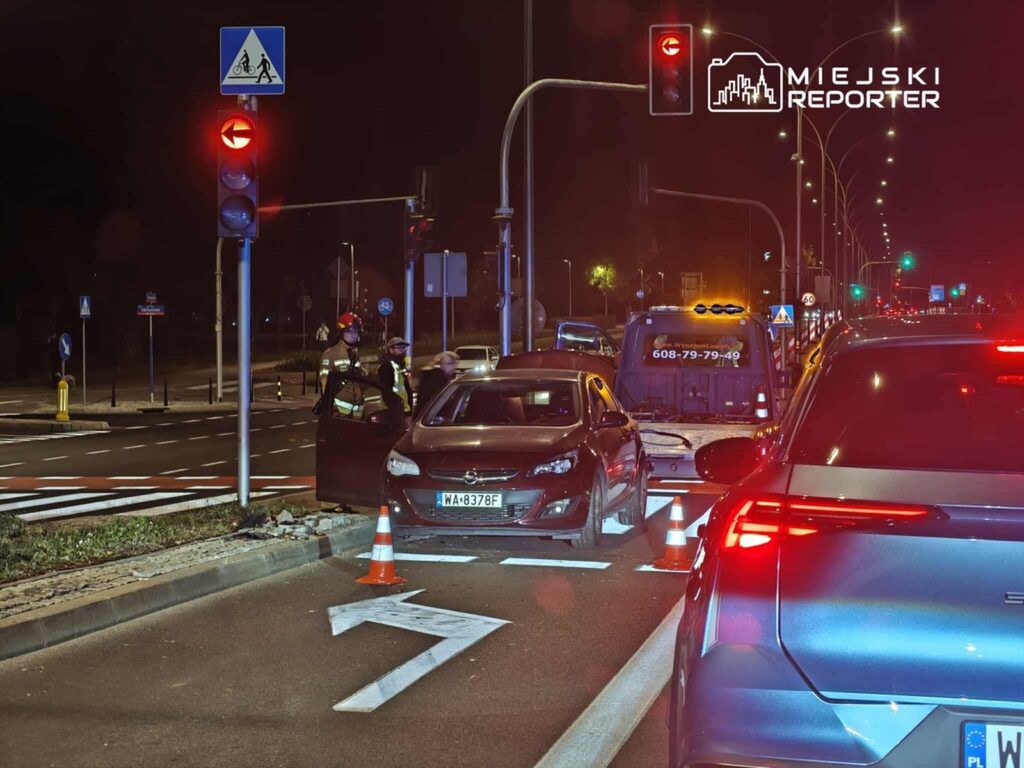 Policjanci rozmawiają z kierowcą zatrzymanego samochodu przy skrzyżowaniu, w tle czerwone światło.