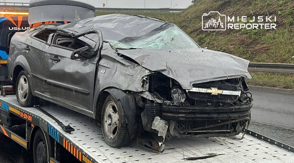 Zniszczony samochód osobowy na lawecie, stojący na poboczu drogi, z wyraźnymi uszkodzeniami przodu i dachu.