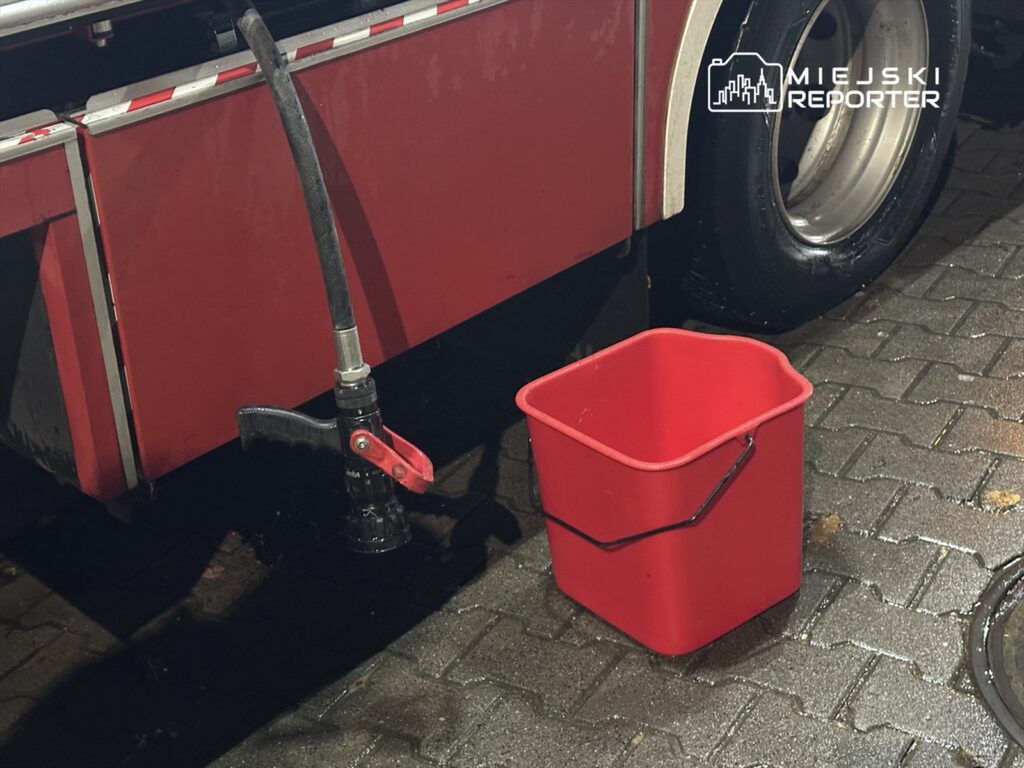 Czarna wąż z dyszą podłączony do czerwonego wozu strażackiego, obok czerwona wiadro na mokrej kostce brukowej.