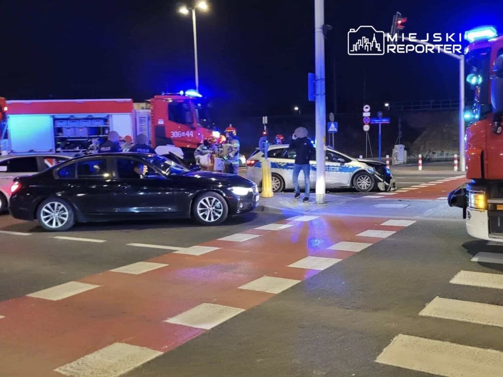 Na skrzyżowaniu nocą, czarne auto stoi obok uszkodzonego pojazdu policji, a ratownicy i policjanci prowadzą działania.