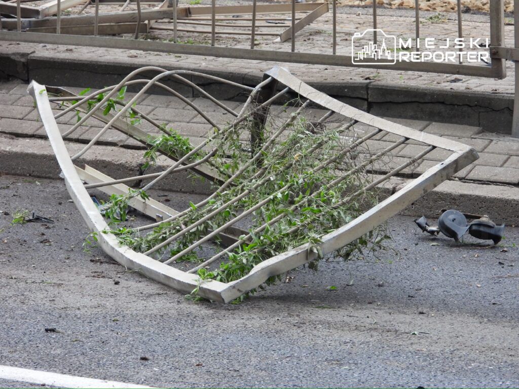 Złamana metalowa ławka z roślinnością leży na ulicy obok chodnika.