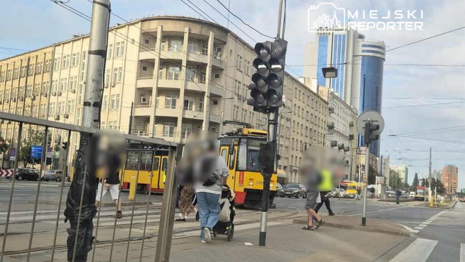 Grupa przechodniów przechodzi przez jezdnię obok tramwaju na przystanku w miejskim otoczeniu. W tle widać nowoczesne budynki.