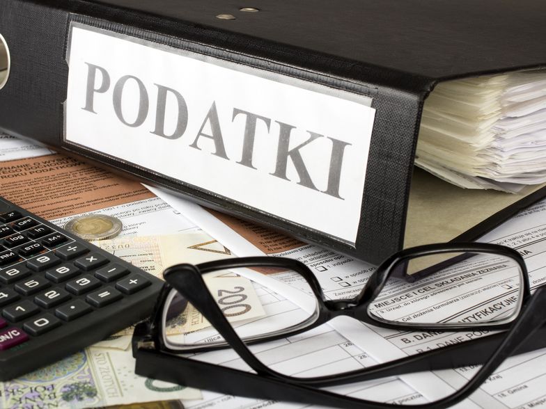 Na zdjęciu formularze podatkowe, na nich leżą okulary, pieniądze i kalkulator. Obok znajduje się segregator z podpisem "podatki"