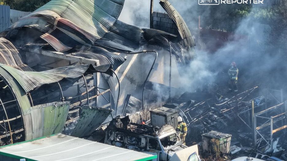 Zniszczony budynek z metalowym dachem, nad którym unosi się dym, w otoczeniu spalonego wraku pojazdu i strażaków gaszących ogień.