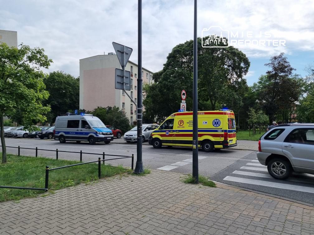 Karetka i radiowóz policyjny zatrzymane na skrzyżowaniu w otoczeniu bloków mieszkalnych.