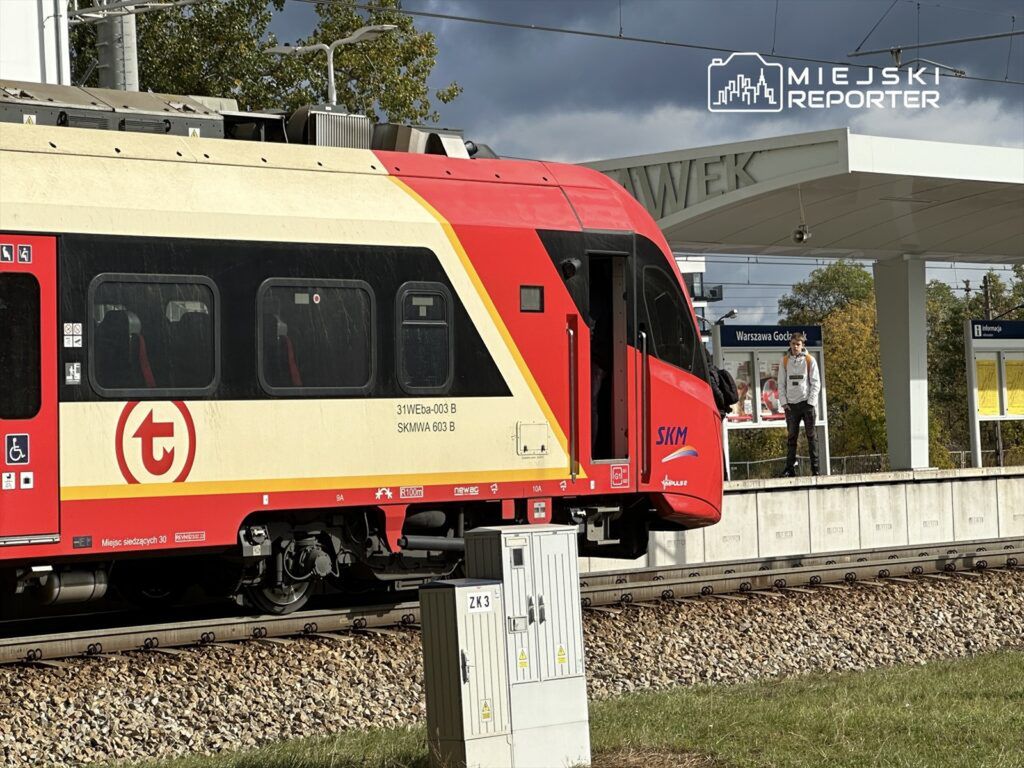 Pociąg SKM zatrzymany na stacji, a młody mężczyzna stoi na peronie obok. W tle widoczny budynek stacyjny.