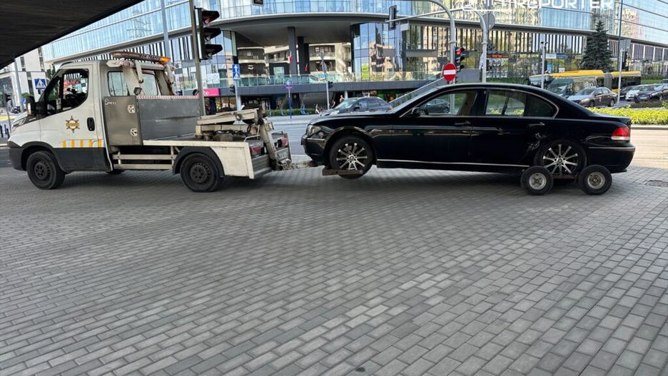 Laweta holująca czarny samochód osobowy na ruchliwej ulicy, obok nowoczesnych budynków i sygnalizacji świetlnej.