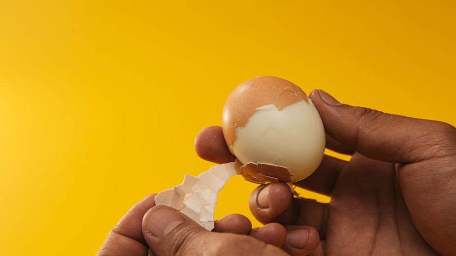Close-up of hands peeling a boiled egg against a bright yellow backdrop, showcasing the texture.