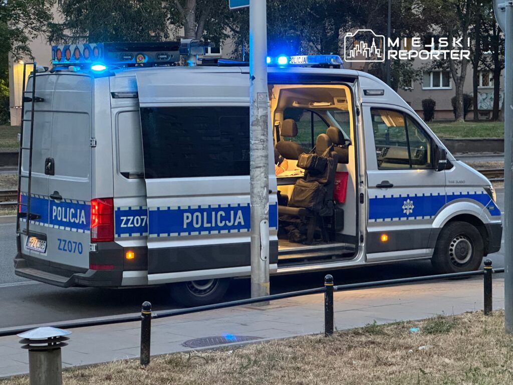 Policyjny van z włączonymi sygnałami świetlnymi, zaparkowany przy ulicy, z otwartymi tylnymi drzwiami.