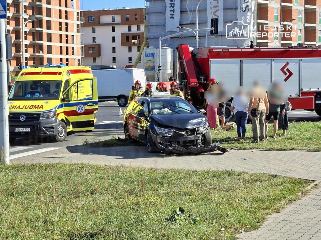 W miejscu zdarzenia stoi czarny samochód osobowy z uszkodzonym przodem, obok niego zespół ratunkowy oraz strażacy rozmawiający z zebranymi osobami.