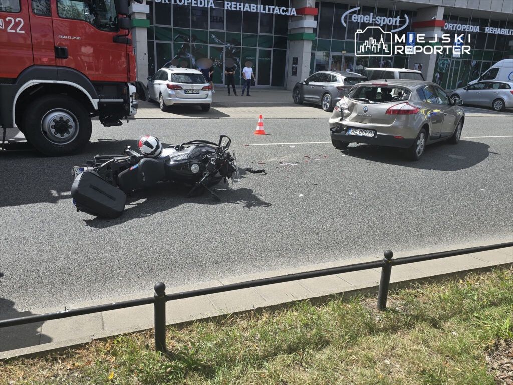 Motocykl leży na jezdni obok uszkodzonego samochodu, a w tle widać budynek z reklamą oraz pojazdy zaparkowane na ulicy.