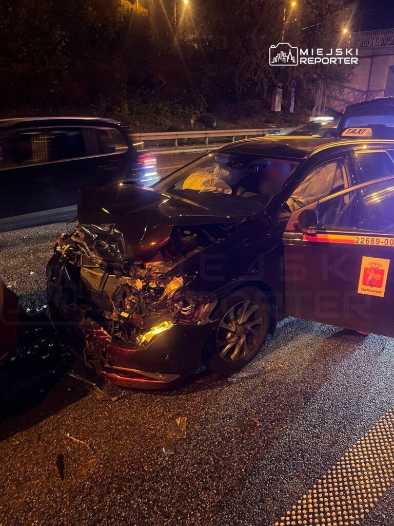 Czarny samochód osobowy po kolizji z uszkodzoną przednią częścią stoi na drodze w nocy. Na jezdni leżą odłamki szkła.