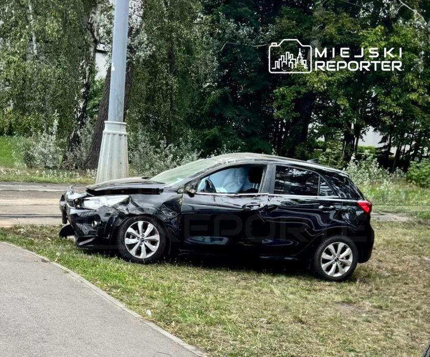 Czarny samochód osobowy z uszkodzoną przednią częścią zaparkowany na trawie obok chodnika w otoczeniu drzew.