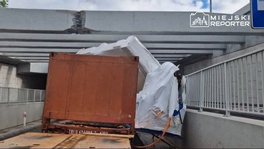 Zniszczony kontener wjeżdżający pod niską estakadę, z widocznym uszkodzeniem dachu konstrukcji.