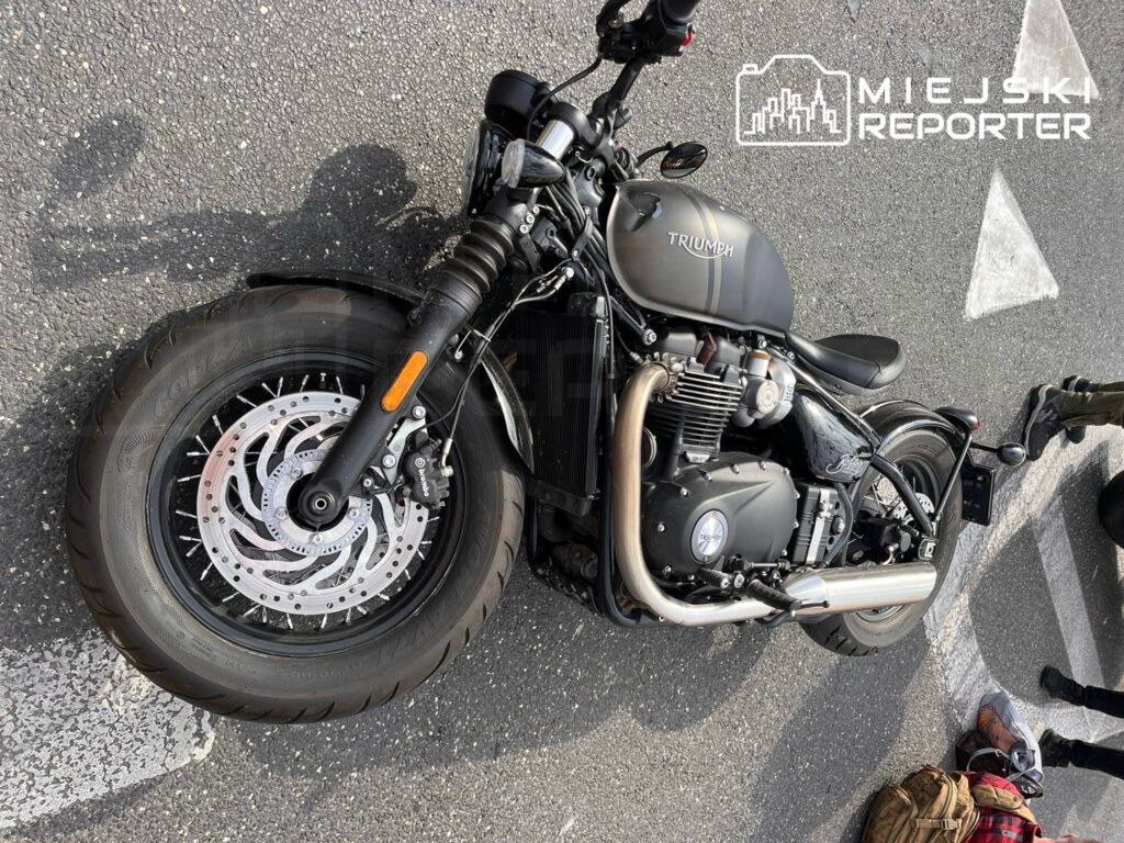 Motocykl marki Triumph leży na asfalcie, częściowo zasłonięty przez torbę i obuwie. W tle widoczni są ludzie, którzy mogą być świadkami zdarzenia. Obraz ilustruje sytuację po ewentualnym wypadku lub awarii jednośladu. Zdarzenie miało miejsce na drodze.