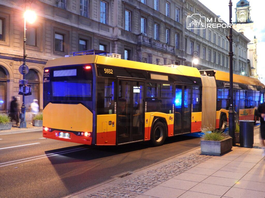 Jasnożółty autobus przegubowy zatrzymany na ulicy w nocy, obok historycznego budynku.