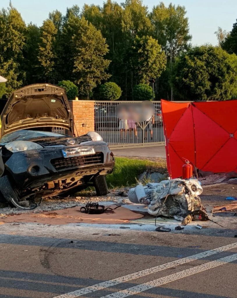 Zniszczony samochód leży na boku obok drogi, obok niego przewrócony silnik i czerwona parasolka ochronna.