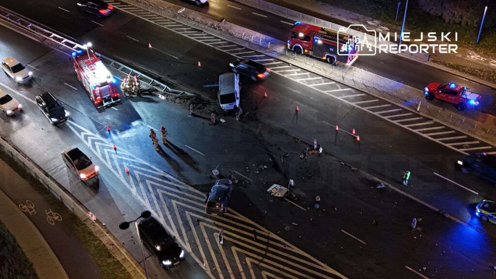 W nocy na skrzyżowaniu ruchliwej drogi strażacy i policjanci zabezpieczają miejsce wypadku, obok leży uszkodzony samochód.