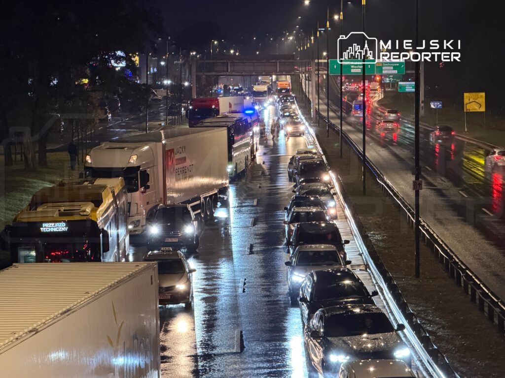 Korki na drodze, pojazdy stojące w deszczu, oświetlone latarniami w nocy, w tle znaki drogowe.