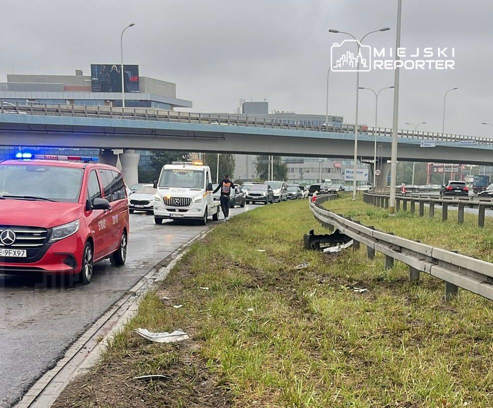 Na drodze stoi czerwony samochód z włączonymi sygnałami świetlnymi, obok niego porusza się pracownik, a w tle widać zator drogowy.