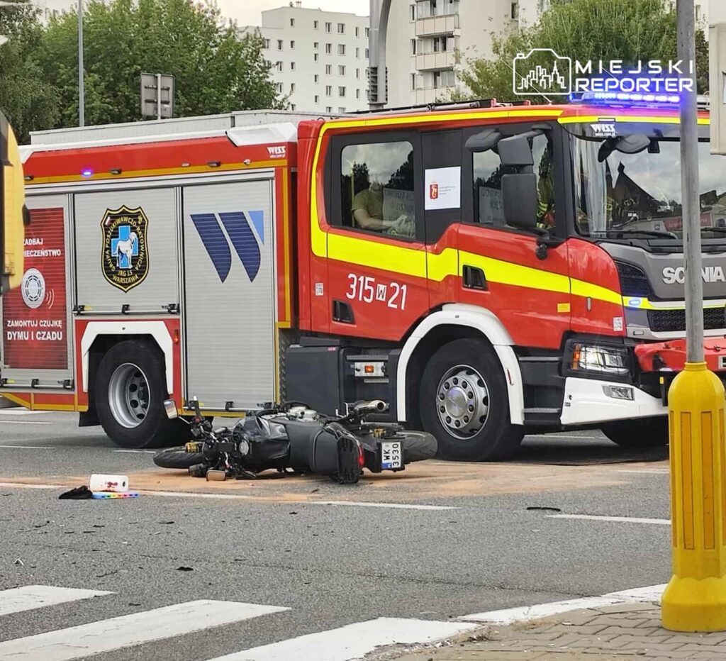 Na zdjęciu widoczny jest wypadek drogowy z udziałem motocykla oraz wozu strażackiego. Motocykl leży na jezdni obok pojazdu strażackiego. W tle widać budynki mieszkalne. Sytuacja wskazuje na interwencję służb ratunkowych, które zajmują się zdarzeniem.