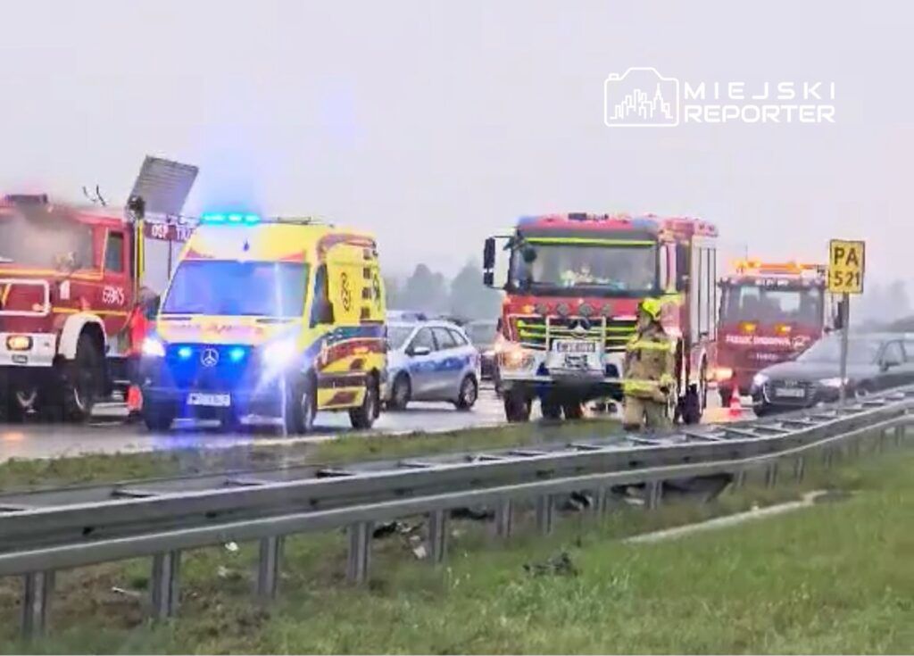 Karetka i samochody straży pożarnej interweniują na drodze, obok stojących pojazdów w deszczowej pogodzie.