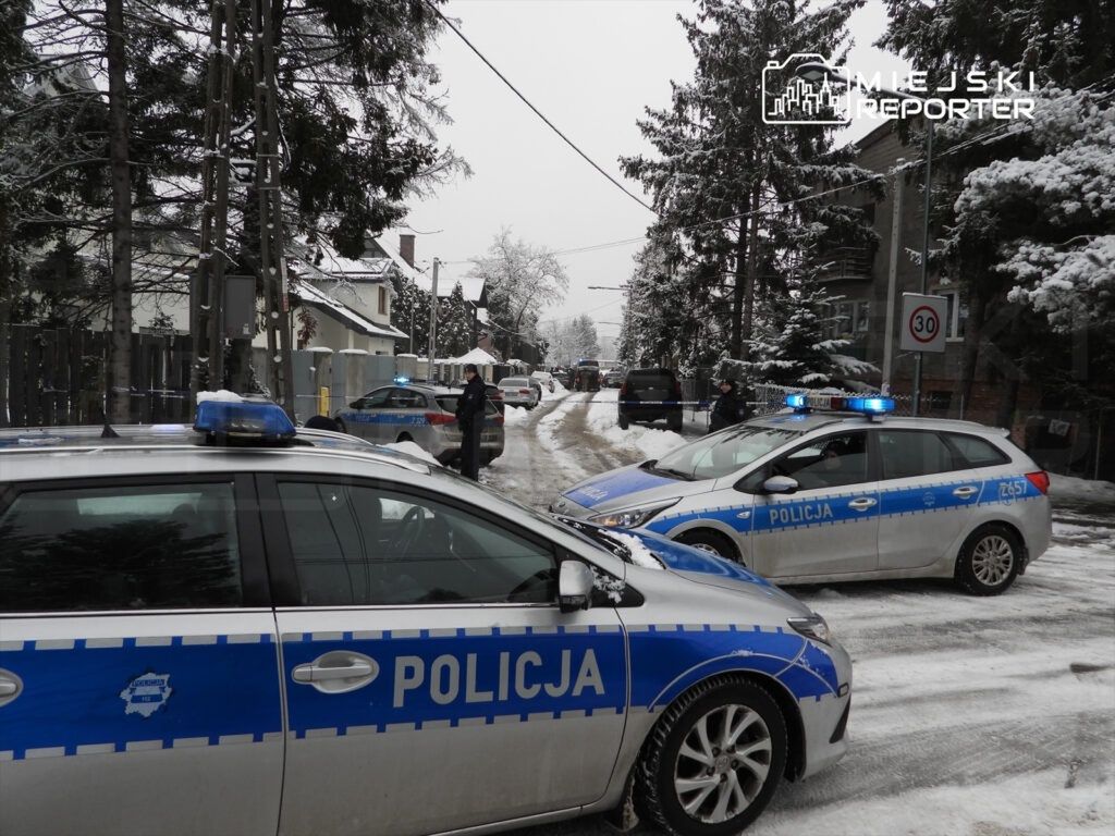 Policyjne radiowozy z włączonymi sygnałami świetlnymi stoją na zaśnieżonej ulicy, a policjanci prowadzą działania.