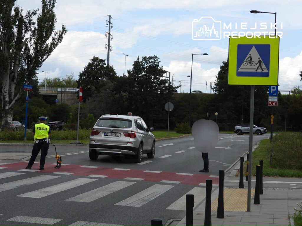 Policjant mierzy odległość na przejściu dla pieszych, obok zatrzymany samochód osobowy. W tle widać znaki drogowe i zieleń.