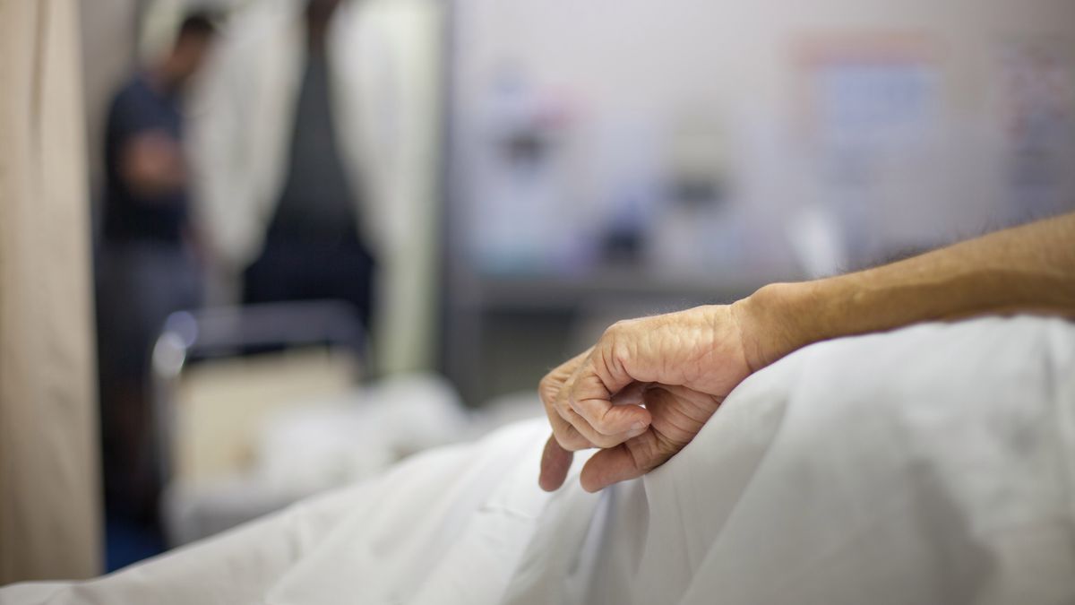 An old man's hand rests at his side in a hospital bed