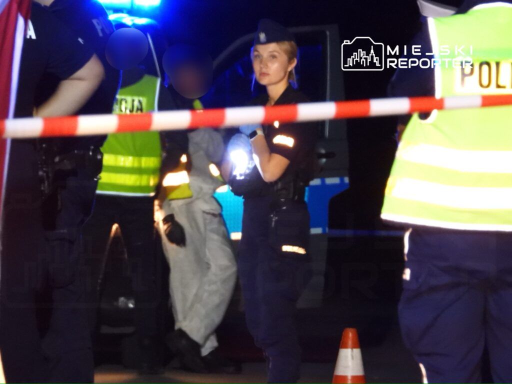 Interwencja policji w nocy, na miejscu zdarzenia. Funkcjonariusze zabezpieczają teren, a w tle widać pojazdy służb ratunkowych. Na pierwszym planie policjantka z latarką, obserwująca sytuację. Wszyscy uczestnicy zdarzenia są w odpowiednich strojach ochronnych.