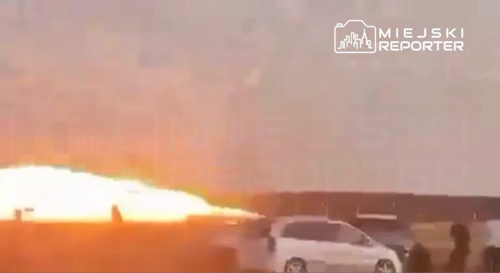 Grupa osób obserwująca wystrzał ognia z pojazdu na otwartej przestrzeni, w tle widać zachód słońca.