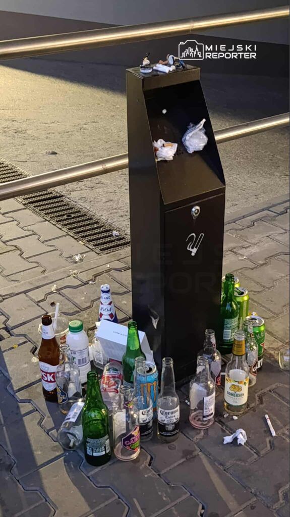 Kosz na odpady zapełniony butelkami i puszkami, otoczony przez śmieci na chodniku w miejskim otoczeniu.