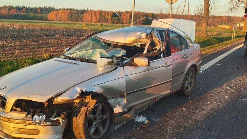 Uszkodzony samochód osobowy leży na poboczu drogi, obok niego stoi wóz strażacki, a w tle widać pola i drzewa.