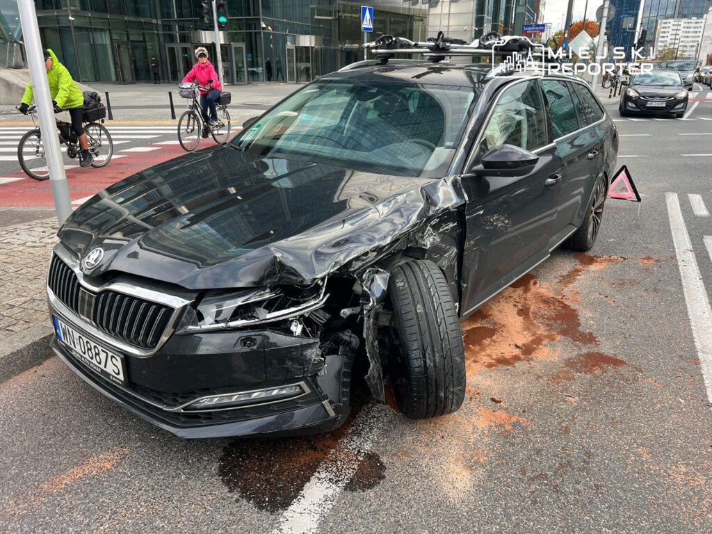 Czarny samochód osobowy z uszkodzoną lewą stroną stoi na ulicy, obok rowerzyści przejeżdżają na tle nowoczesnych budynków.