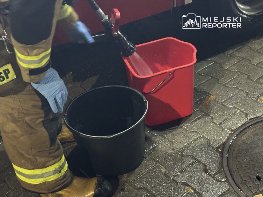 Strażak w mundurze i rękawiczkach napełnia czerwony pojemnik wodą z węża, obok czarny wiadro na mokrej kostce brukowej.