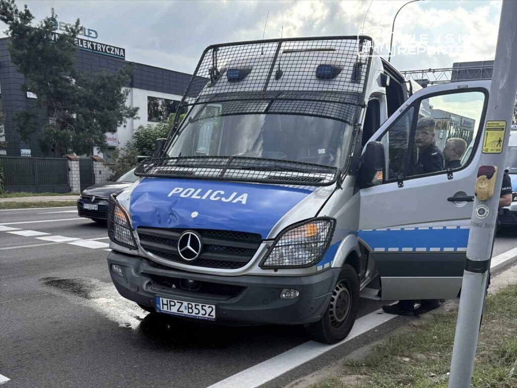 Policyjny furgon z uszkodzoną przednią częścią zatrzymany na ulicy, obok widać przechodnia i sygnalizator.
