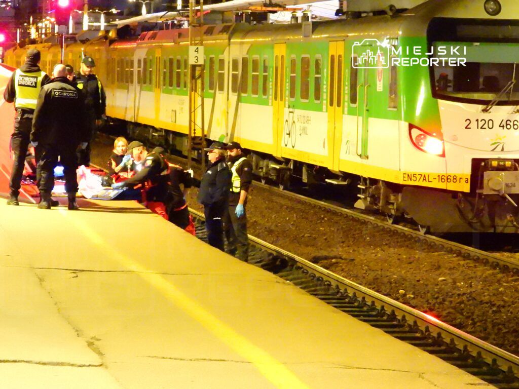 Grupa ratowników medycznych udziela pomocy poszkodowanemu na peronie, obok zatrzymanego pociągu.