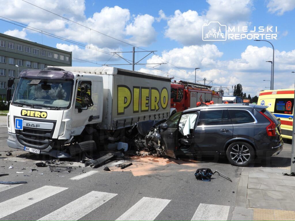 Zderzenie ciężarówki z osobowym samochodem na skrzyżowaniu, wokół obecne służby ratunkowe oraz uszkodzone pojazdy.