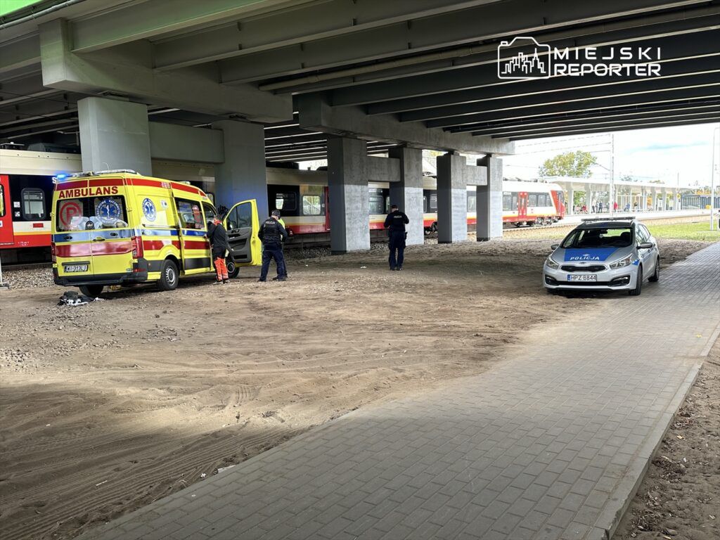 Karetka pogotowia oraz radiowóz policji stoją pod estakadą, a funkcjonariusze rozmawiają przy otwartych drzwiach ambulansu.