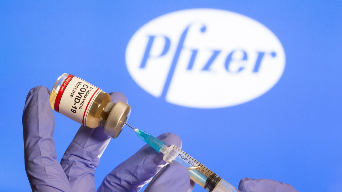 A woman holds a small bottle labeled with a "Coronavirus COVID-19 Vaccine" sticker and a medical syringe in front of displayed Pfizer logo in this illustration taken, October 30, 2020.